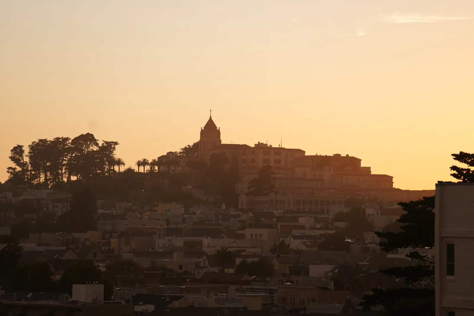 A golden sunset over a city hillside with a cathedral tower silhouetted above rooftops and trees.