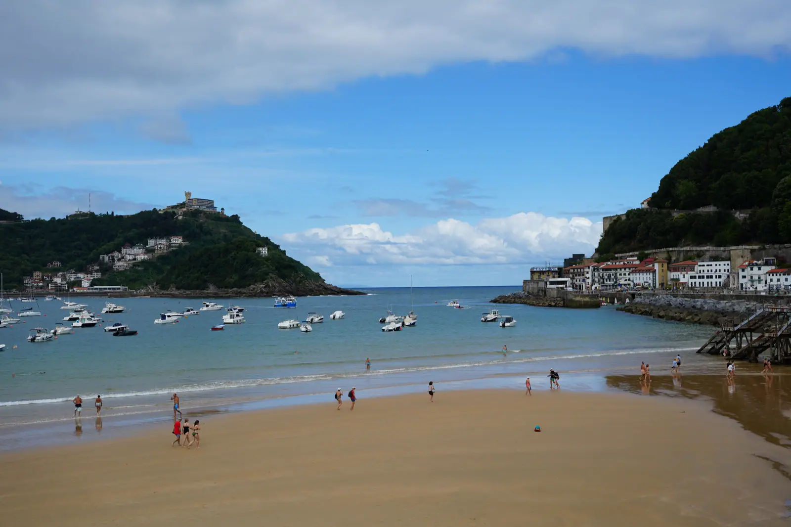 A sandy beach beside a calm bay with several small boats, people walking near the water, and green hills under a partly cloudy sky.