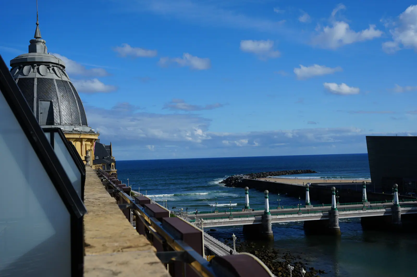 A domed building beside a coastal bridge and beach under a blue sky.