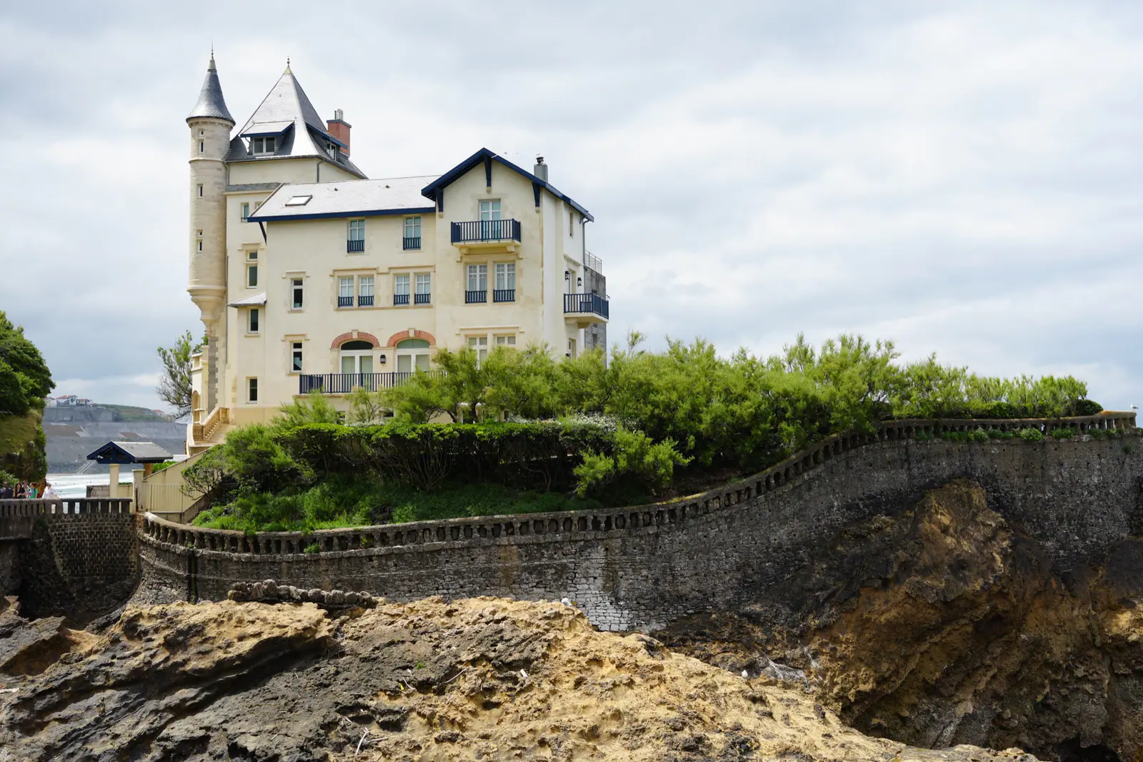A cream-colored seaside villa with a turret sits above rocky cliffs and a stone wall, with ocean visible to the left.