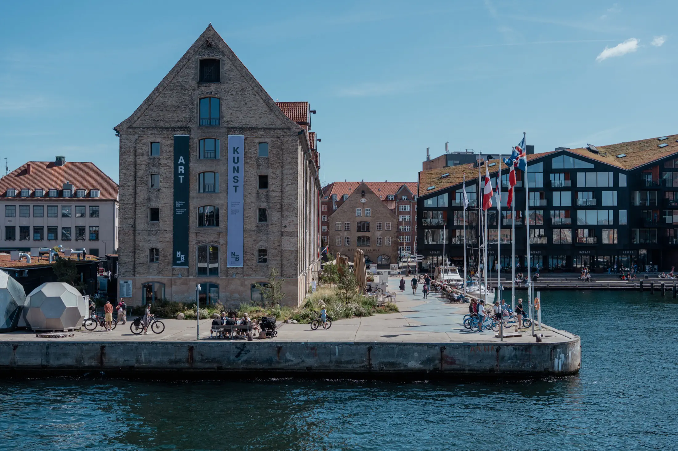 Waterfront buildings along a canal with people walking and cycling on the quay, flags on poles, and a historic brick building labeled Kunst.