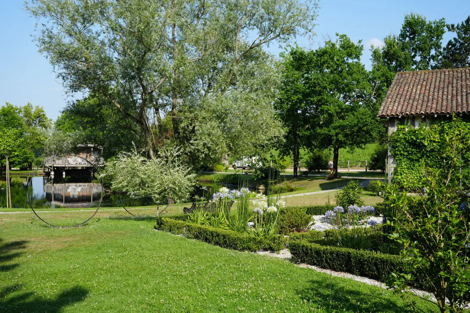 Sunny landscaped garden with a pond, trees, flowering plants, and a rustic tiled-roof building.