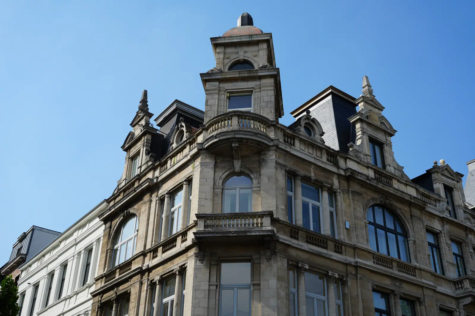 Low-angle view of an ornate historic stone building with balconies, arched windows, and dormer roofs against a blue sky.