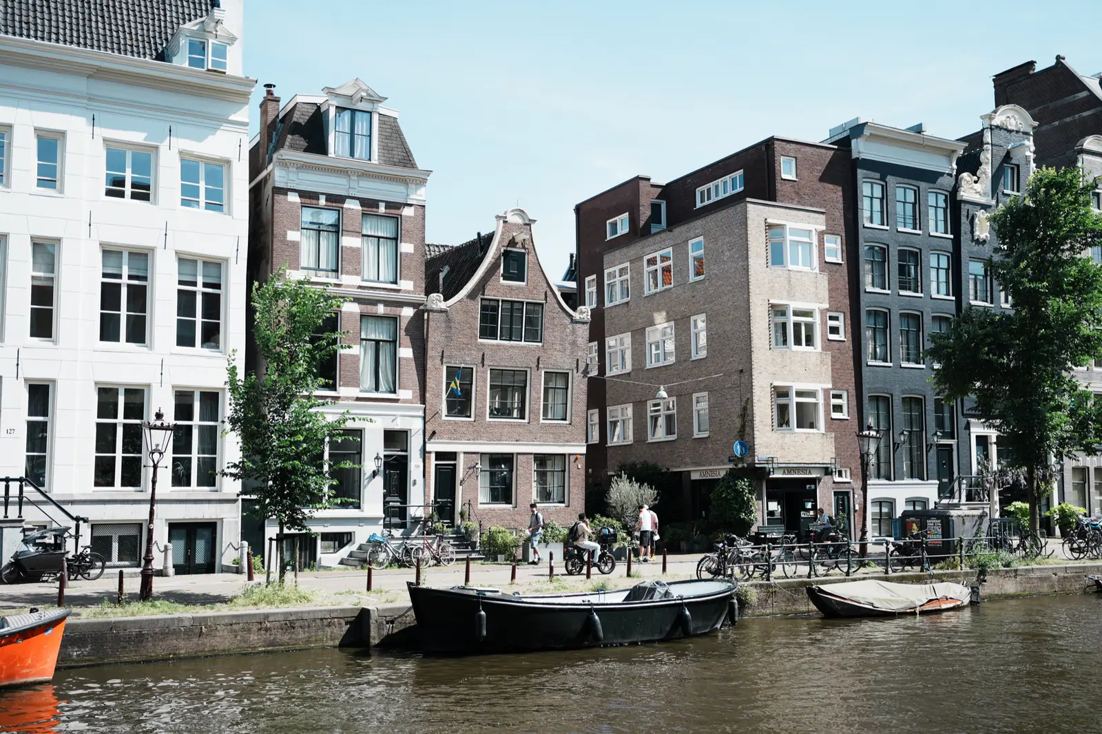Canal-side street in Amsterdam with historic buildings, bicycles, pedestrians, and boats moored along the water.