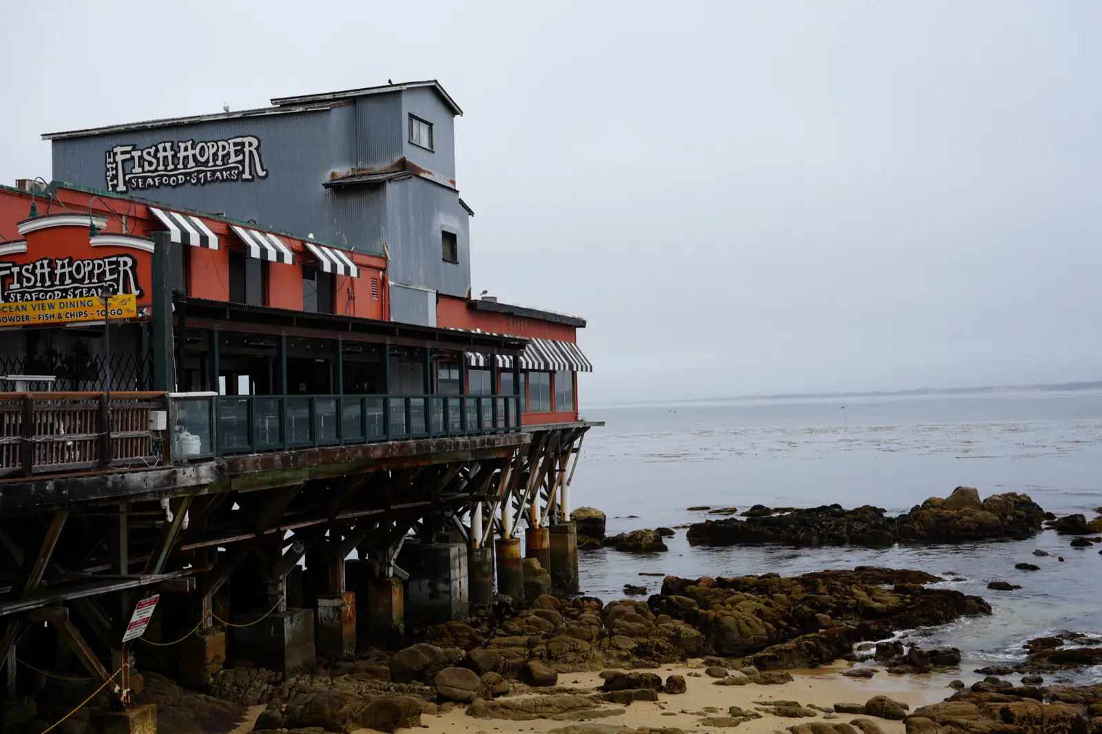 Seaside restaurant on pilings above a rocky shoreline with calm water and an overcast sky.