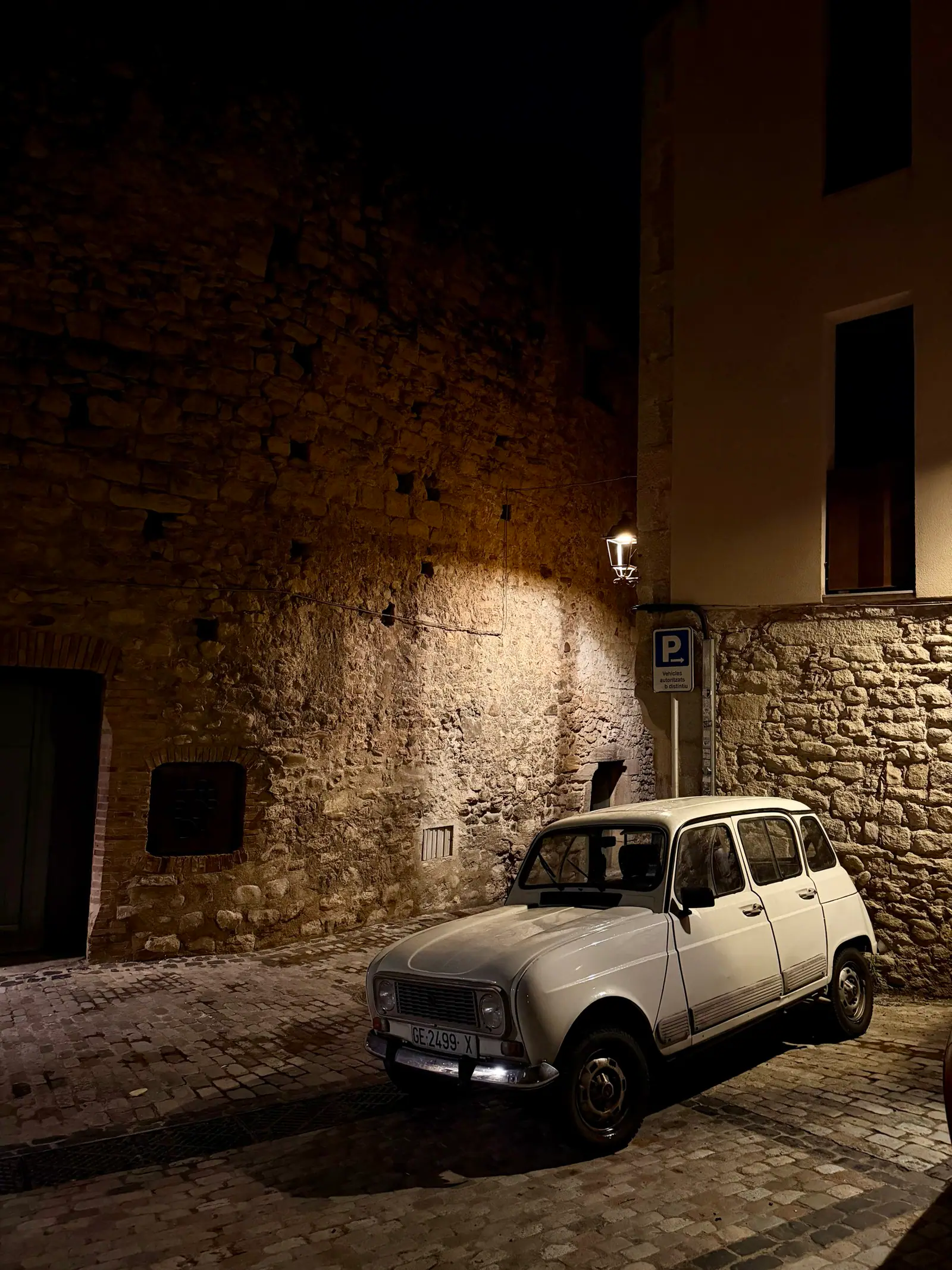 A narrow cobblestone street at night with stone walls, a parked white car, and a lit building facade.
