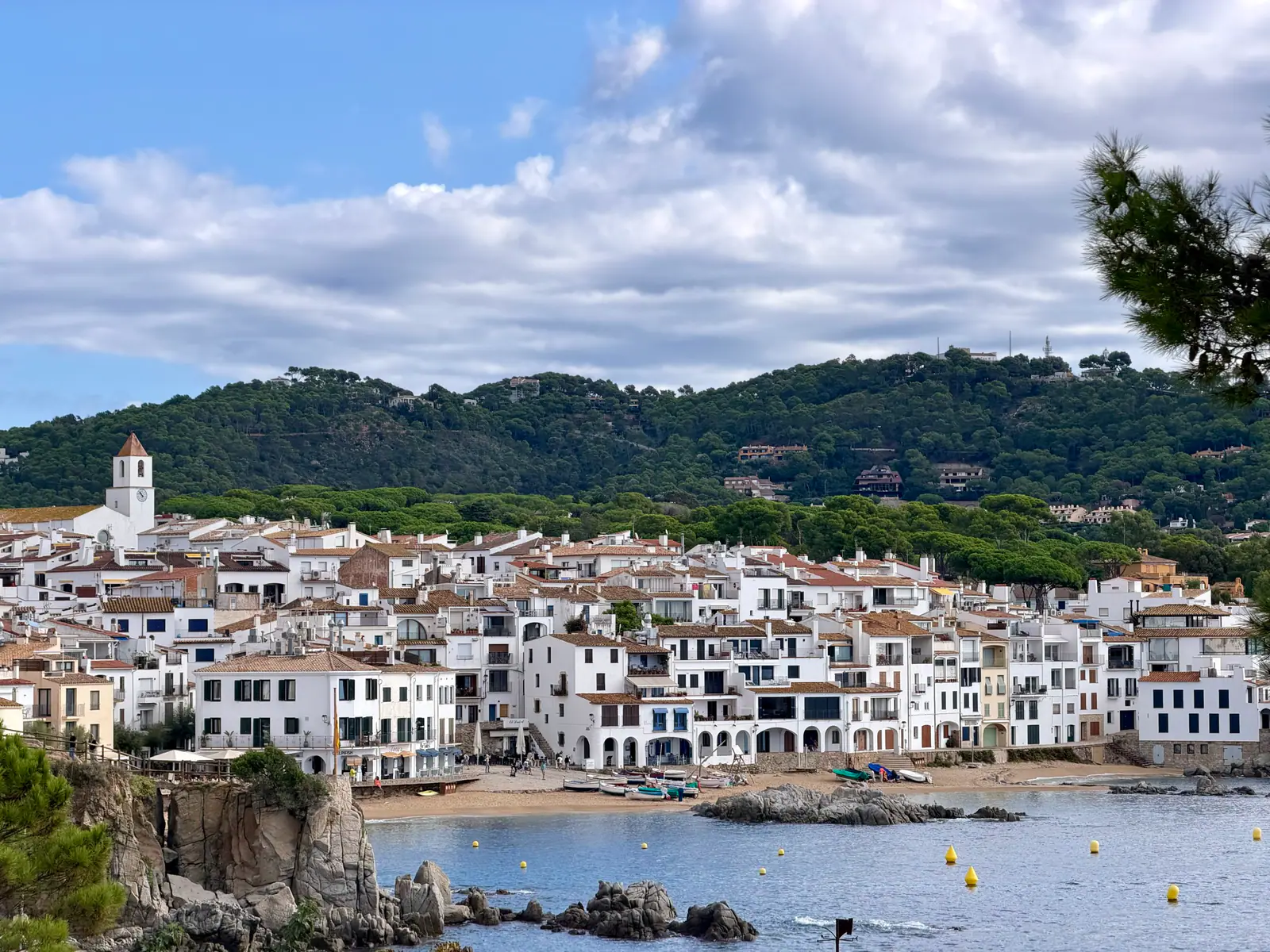 A white seaside village beside a calm bay with a church tower, rocky shore, and green hillside behind it.
