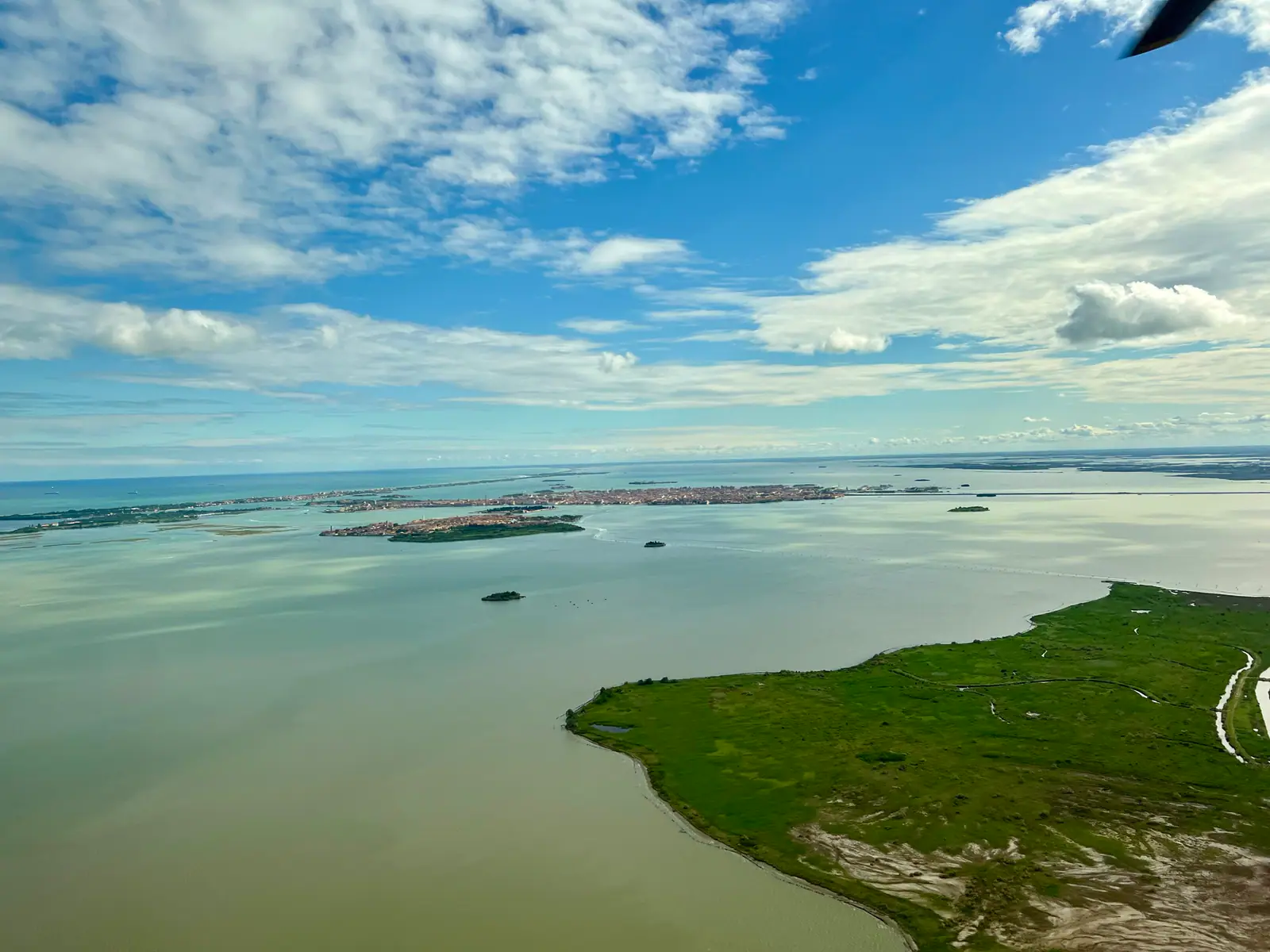 Aerial view of the Venice lagoon with islands, marshland, and open water beneath a cloud-filled blue sky.